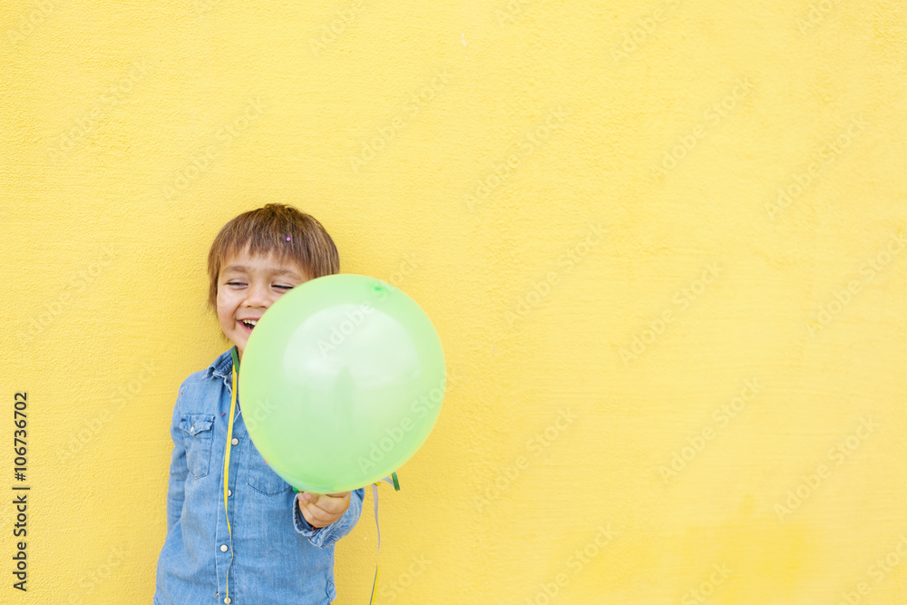 Smiling little boy with green balloon and streamer standing in front of yellow wall Smiling little boy with green balloon and streamer standing in front of yellow wall