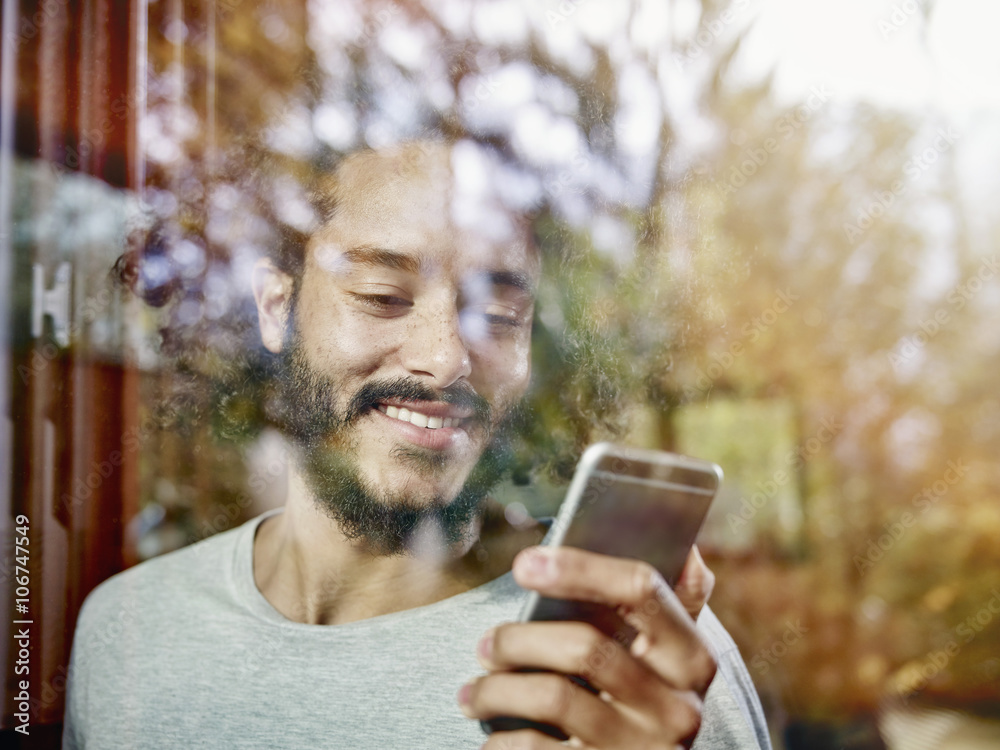 Smiling young man looking on cell phone behind windowpane Smiling young man looking on cell phone behind windowpane