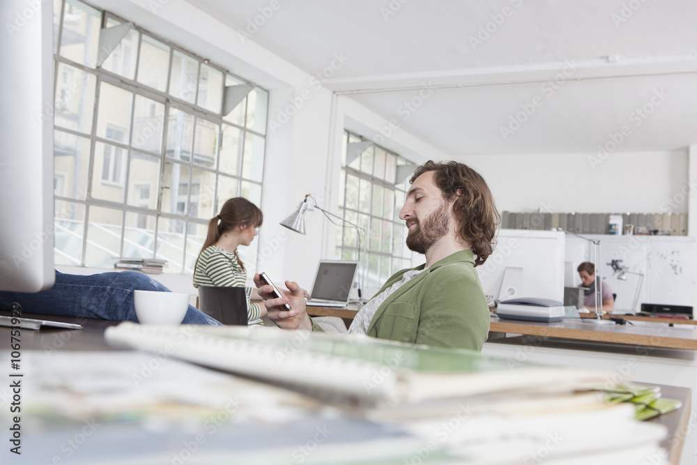 Young man sitting at his desk in a creative office using his smartphone Young man sitting at his desk in a creative office using his smartphone