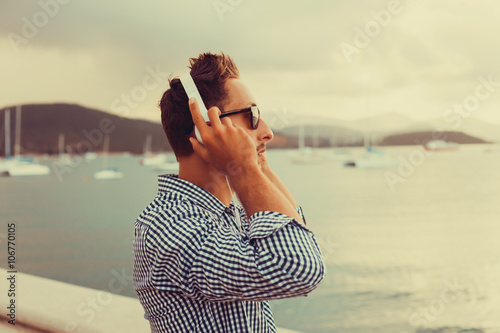 stylish young man in a shirt and shorts and sneakers listening to music in headphones on a smartphone and is sitting on pier looking at sea on yachts boats and the sunset,music lounge ,relaxing 