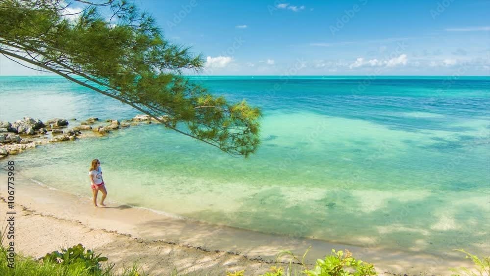 A Young Woman Walking Alone on a Tropical Beach in Bermuda with Golden Sand and Turquoise Water ending on the Horizon with a Blue Sky and White Clouds. Ultra High Definition, 4K