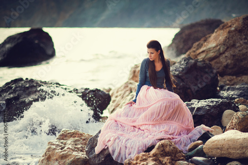 A girl sits on a rock on the beach against the sky and the sea, lifestyle, recreation, solitude, meditation