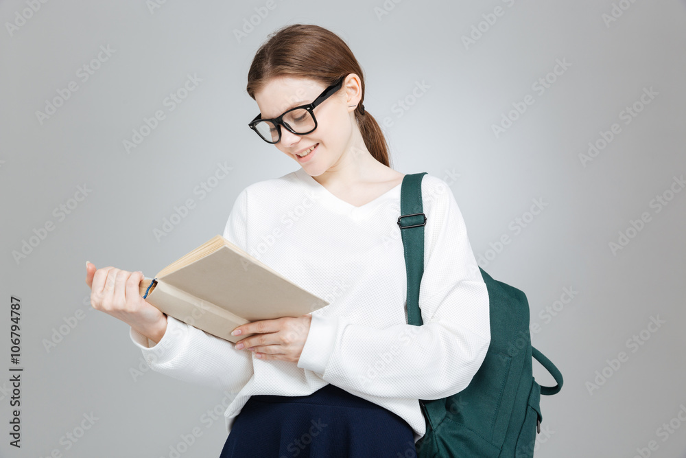 Cheerful lovely teenage girl in glasses with backpack reading book