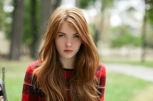 Closeup portrait of young adorable redhead woman in red plaid jacket stares into camera with blurred park background photo