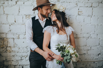 girl with a wreath of white flowers on the head in a white wedding dress and a bearded man in a suit and a hat are holding a bouquet of white flowers and green against a white brick wall