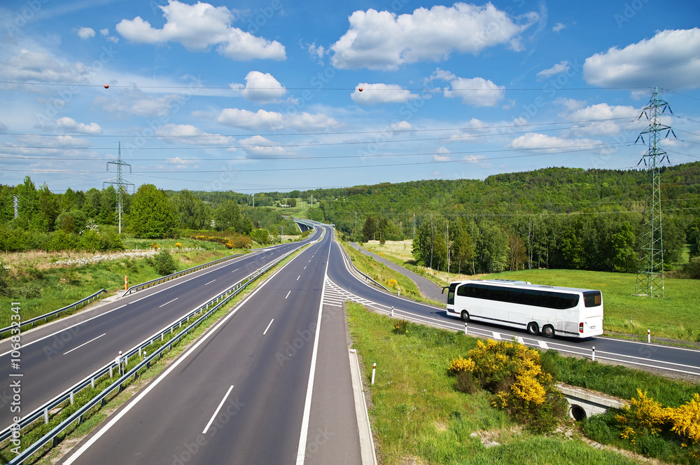 White Bus entering on an empty asphalt highway in the landscape. High power line pylons leading across the freeway. Sunny summer day with blue skies and white clouds. View from above.