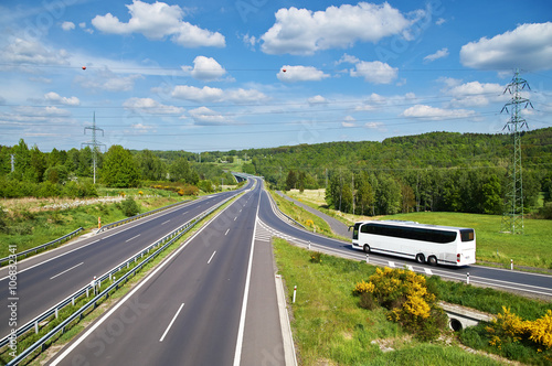 Obraz na płótnie White Bus entering on an empty asphalt highway in the landscape. High power line pylons leading across the freeway. Sunny summer day with blue skies and white clouds. View from above. w salonie