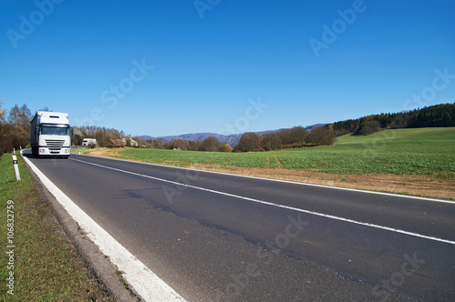 White trucks arriving on the asphalt road from afar in the countryside in early spring. Flowering trees. Wooded mountains in the background. Sunny day with blue skies.
