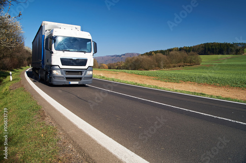 White truck arriving on asphalt road in a rural landscape in early spring. Flowering trees. Wooded mountains in the background. Sunny day with blue skies.