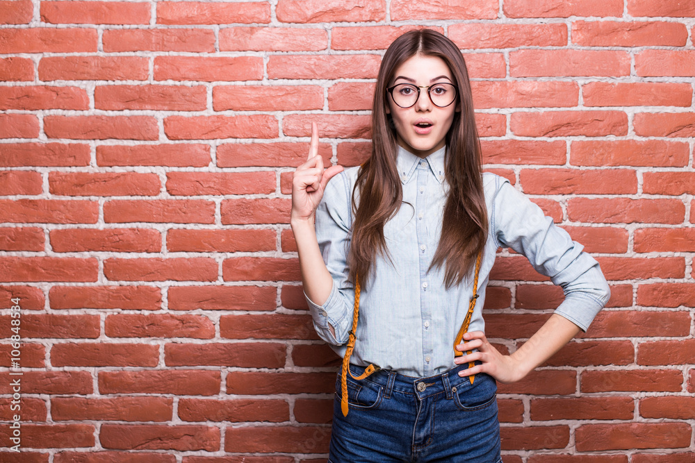 Portrait of beautiful girl in casual clothes showing UP sign, looking in camera and smiling while standing against red brick wall