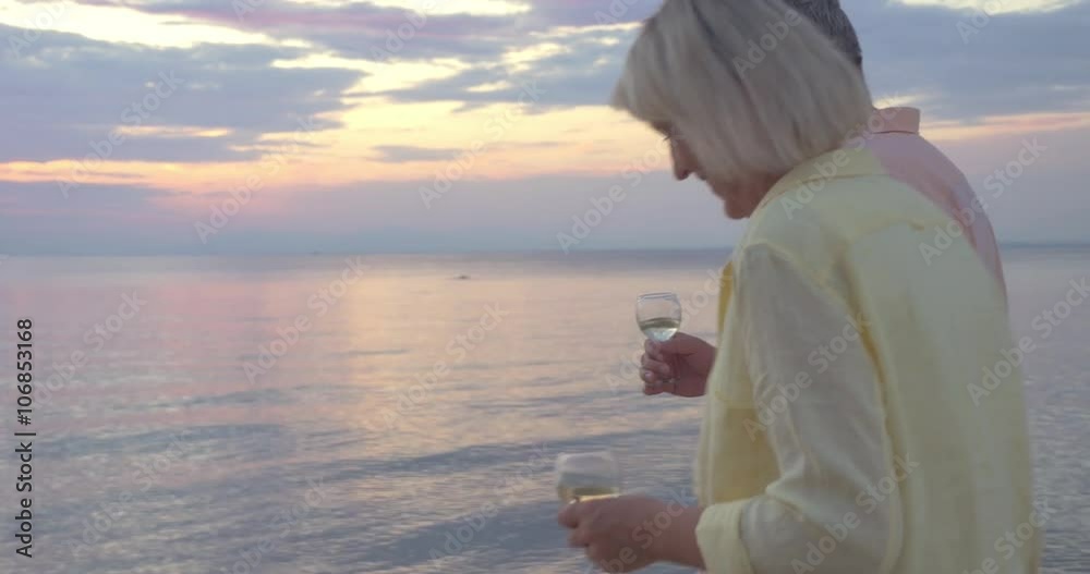 Steadicam shot of mature couple walking by the sea at sunset with wine glasses in hands. Steadicam shot of mature couple walking by the sea at sunset with wine glasses in hands.