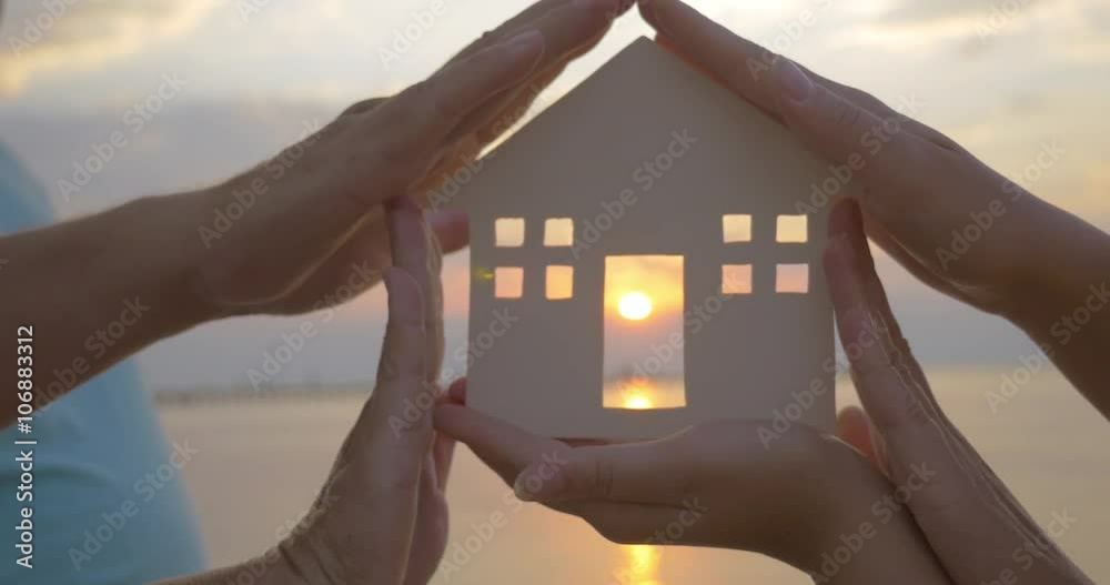 Closeup shot of hands holding house silhouette. Setting sun is seen through the house door.
