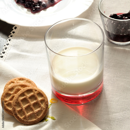 colazione con biscotti, un bicchiere di latte e frutti di bosco, su una tovaglietta ricamata, illuminata dal sole, formato quadrato photo