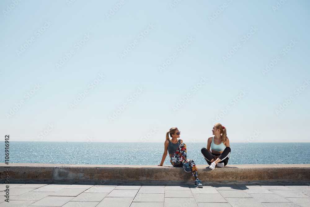 Sport fitness healthy girls relaxing after training outdoor. Young couple talking sitting in waterfront after running and training exercise outside in summer. Caucasian and asian sports women models.