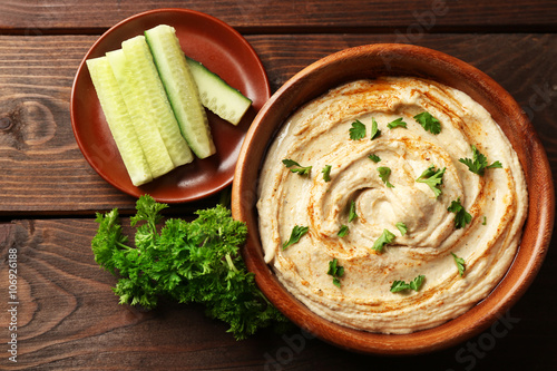 Wooden bowl of tasty hummus with parsley and cucumber on table, close up