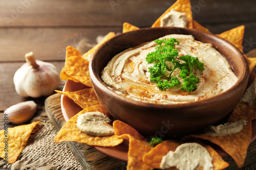 Wooden bowl of tasty hummus with chips, parsley and garlic on table