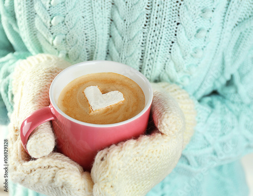 Female hands in warm mittens holding cup of hot cappuccino with heart marshmallow, close up