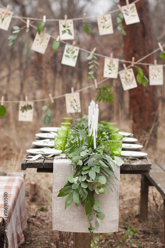 Weddind table setting with white plates and green glasses decorated with white candles, green leaves and eucalyptus and a garland of green leaves and postcards outdoors