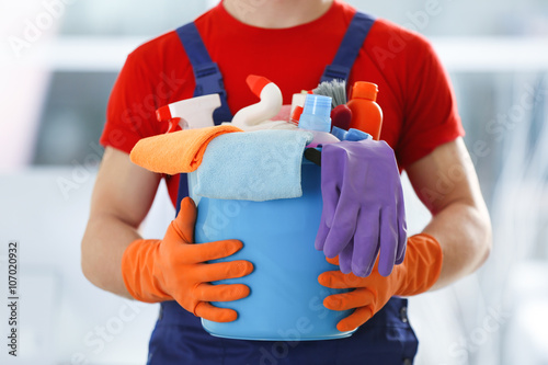 Young janitor holding cleaning products and tools on bucket, close up