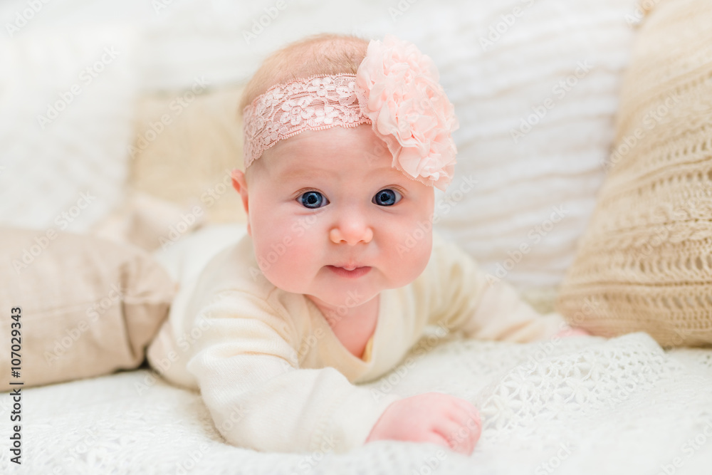 Sweet little baby with chubby cheeks and big blue eyes wearing white clothes and pink band with flower lying on bed and looking at camera. Babyhood and childhood concept Sweet little baby with chubby cheeks and big blue eyes wearing white clothes and pink band with flower lying on bed and looking at camera. Babyhood and childhood concept