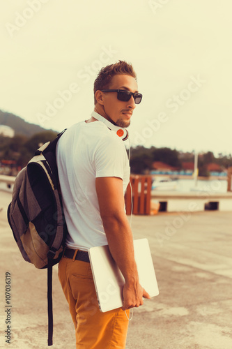 young guy in sunglasses t-shirt holding a working laptop is on the pier with a backpack on shoulders, coolest style,walking away,street style,he works on his laptop to get all his business done early 