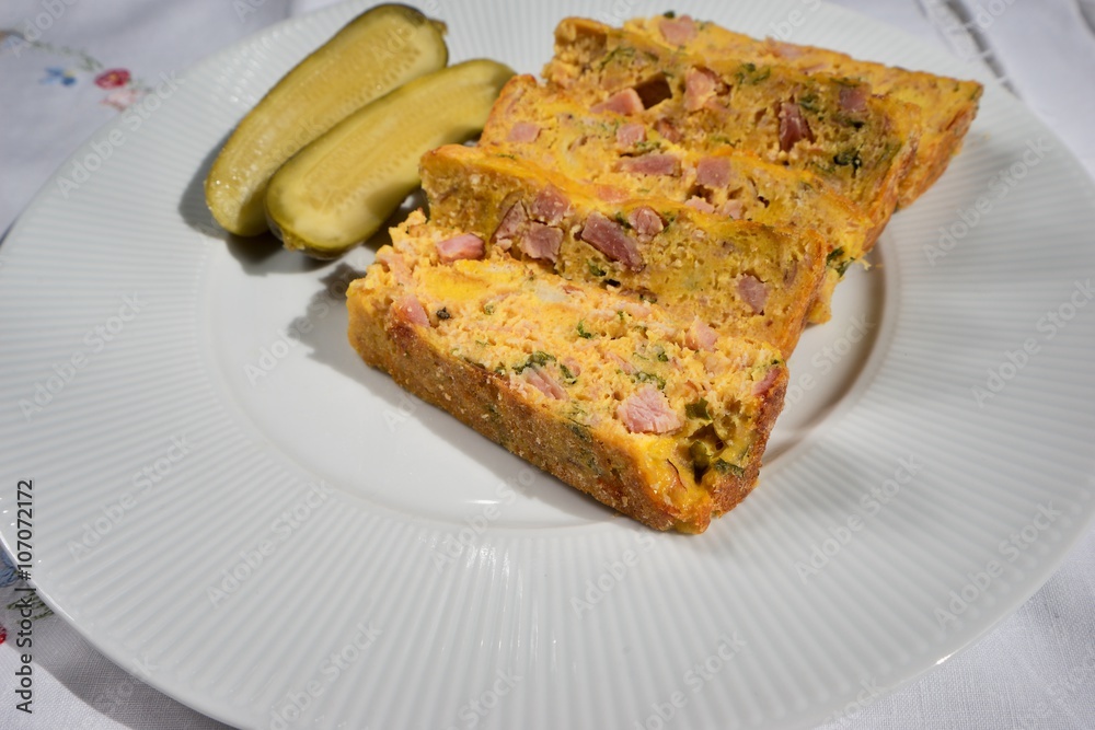 Baked or roasted egg pie or kind of quiche, french style snack servedwhite plate with pickled cucumber. Made from eggs, smoked ham, parsley, pork meat and bread. Traditional food in Czech Republic.