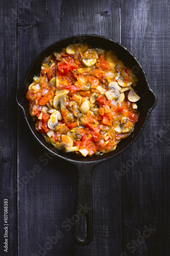 fried mushrooms with vegetables in a black frying pan on a wooden dark background. top view