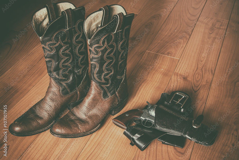 Western Wear Boots and Gun in Holster. Pair of old western cowboy boots and  a revolver in a holster sitting on a hardwood floor. Photos | Adobe Stock