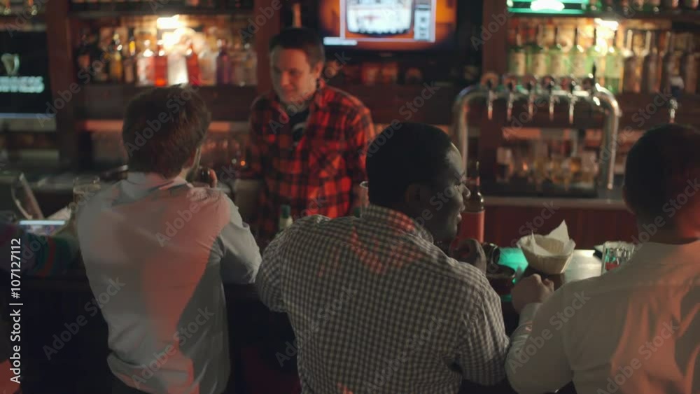 Rear view of men standing in the pub and drinking beer: two of them talking to bartender while three businessmen standing next to them discussing something Rear view of men standing in the pub and drinking beer: two of them talking to bartender while three businessmen standing next to them discussing something