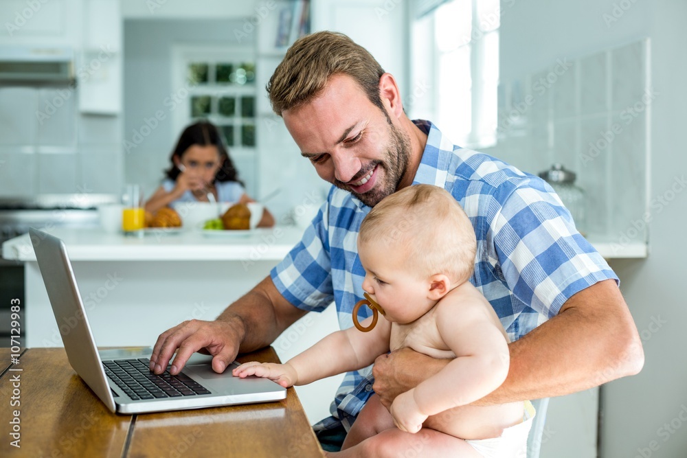 Father looking at baby boy while using laptop at home Father looking at baby boy while using laptop at home