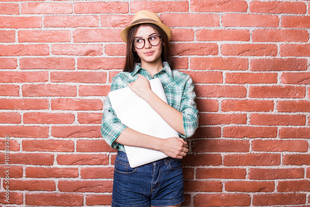 Beautiful young dark-haired girl in casual clothes, hat and eyeglasses posing, smiling and with close laptop in hands, standing against brick wall Beautiful young dark-haired girl in casual clothes, hat and eyeglasses posing, smiling and with close laptop in hands, standing against brick wall