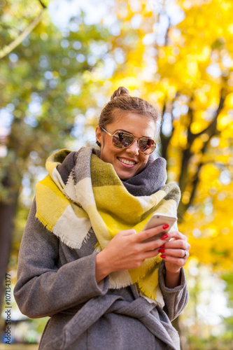 Beautiful young girl using smartphone in park. Autumn time.