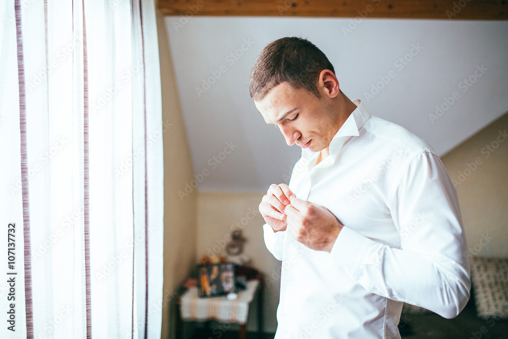 Stylish and handsome groom in waistcoat tying his bow-tie in the morning of the wedding day.