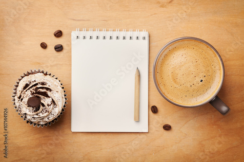 Obraz na płótnie Coffee mug with cupcake, notebook and pencil on rustic table from above, cozy an