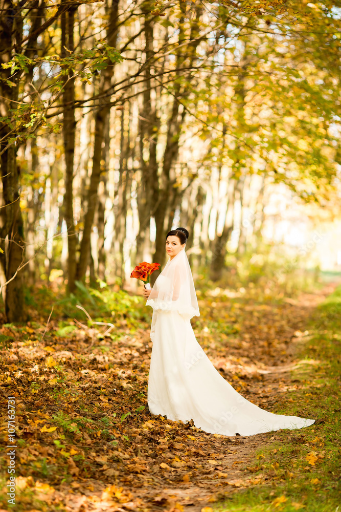 Happy beautiful brunette bride on autumn forest with bouquet of red flowers