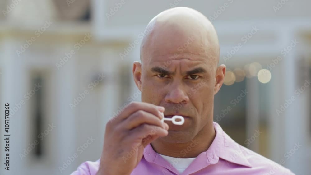 Close up of a mixed-race man in a pink shirt, scowling and blowing bubbles while outdoors on a sunny spring day.  Slow motion, recorded at 180fps
