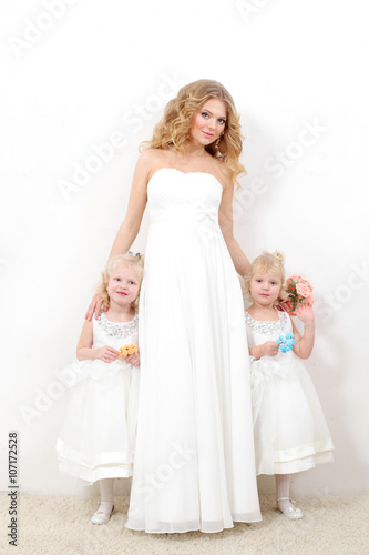 Beautiful blond mom with two young daughters blondes in white dresses on a white background. Beautiful mother and daughters in studio