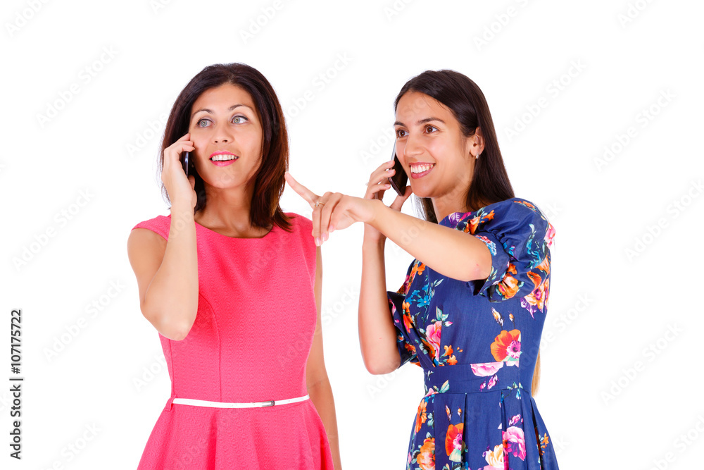 Two beautiful women, girlfriends, sisters in summer colored dress, pointing by finger and speak on mobile cell smartphone on a white background Two beautiful women, girlfriends, sisters in summer colored dress, pointing by finger and speak on mobile cell smartphone on a white background