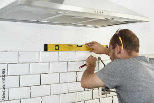 A man working in a new kitchen, a tiler applying tiles to the wall behind the cooker. 