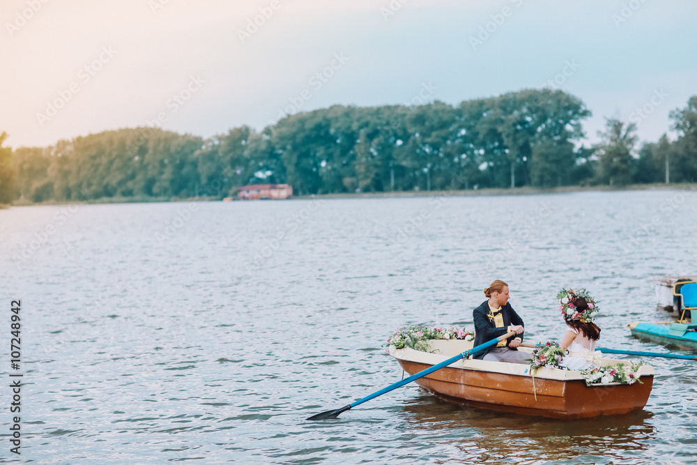 stylish gorgeous happy brunette bride and elegant groom in the boat on the background of a lake. Bride with wreath of flowers