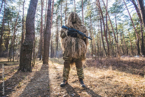 Sniper in camouflaged ghillie suit/Sniper in camouflaged ghillie suit standing in forest with rifle in hands.Low angle view.Selective focus.