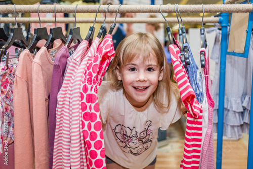 Smiling little girl looks through hangers with clothes. Child has fun in shopping centre.