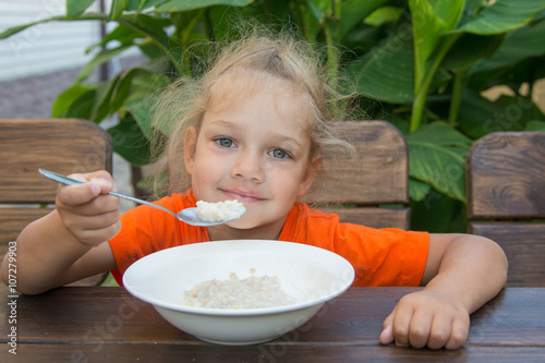 Little girl smiling at a wooden table on a background of green eating porridge