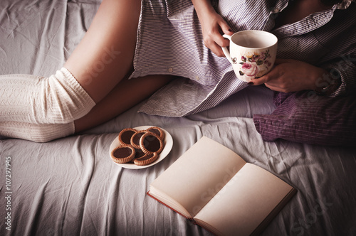 Beautiful sensual young woman reading book and relaxing in bed holding coffe cup in her hand