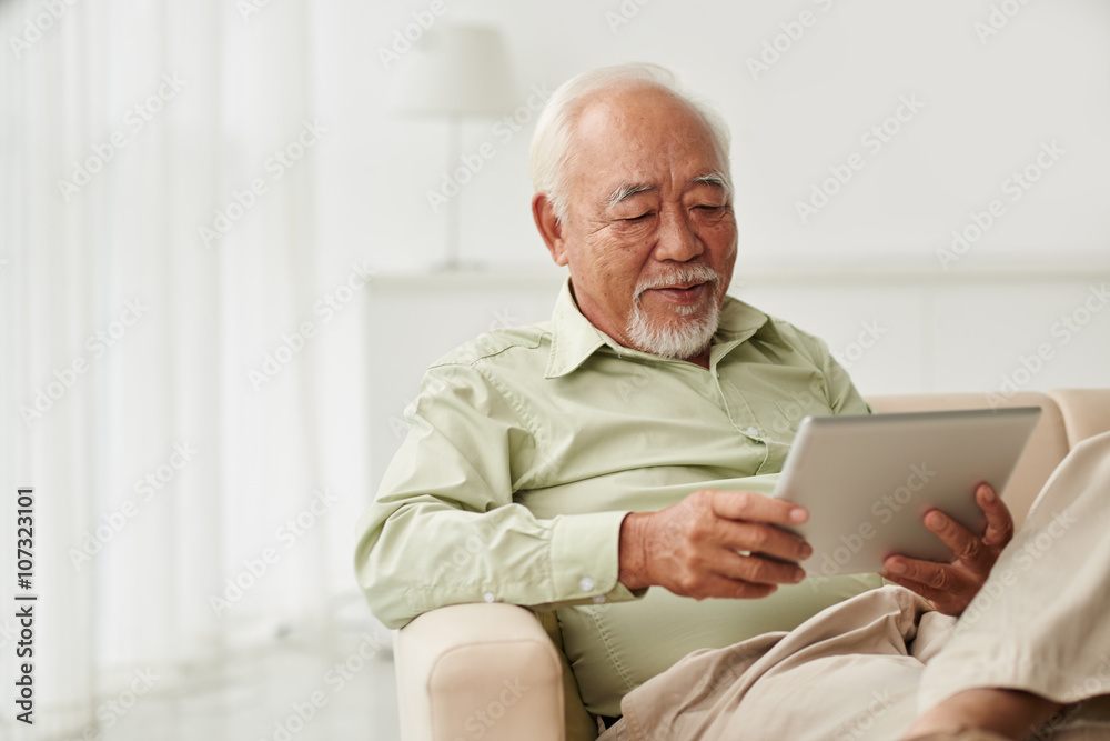 Senior Vietnamese man sitting on sofa and reading new on his tablet computer Senior Vietnamese man sitting on sofa and reading new on his tablet computer