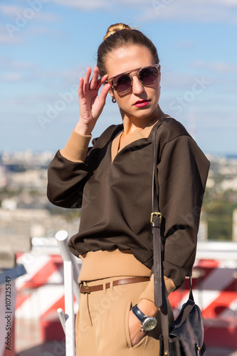 Street fashion portrait stylish pretty woman with sunglasses and bag posing in city on evening sunny sunset 