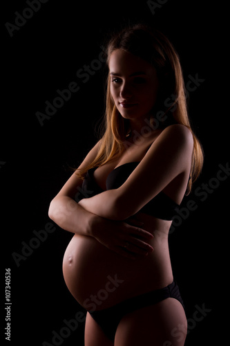 Happy young pregnant woman in black underwear is hugging self on black background © Monstar Studio