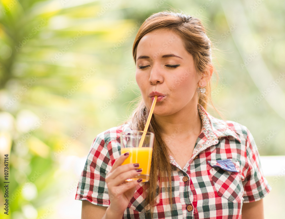 Young woman in square pattern shirt enjoying a yellow juice with eyes closed, garden environment Young woman in square pattern shirt enjoying a yellow juice with eyes closed, garden environment