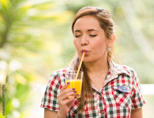 Young woman in square pattern shirt enjoying a yellow juice with eyes closed, garden environment