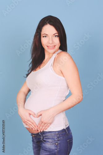 Smiling pregnant woman making heart on tummy over blue. Looking at camera. Maternity. Motherhood. © morrowlight